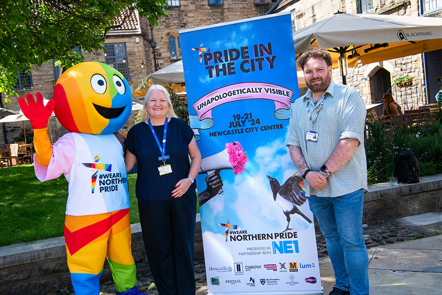 Two Lumo staff members standing with the pride mascot next to a pride in the city poster